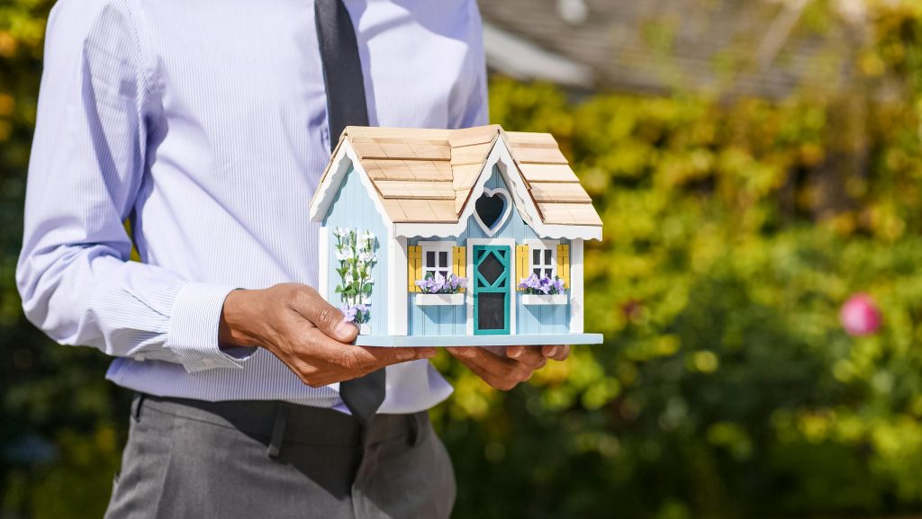 Businessman holds small house model outdoors, symbolizing real estate investment. Un homme d'affaires tient un petit modèle de maison en plein air, symbolisant l'investissement immobilier.